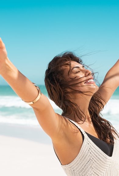 Portrait of healthy young woman standing on the beach with outstretched arms and feeling the breeze. Happy young woman feeling fresh and relaxing at ocean. Latin tanned woman with closedd eyes feeling good.